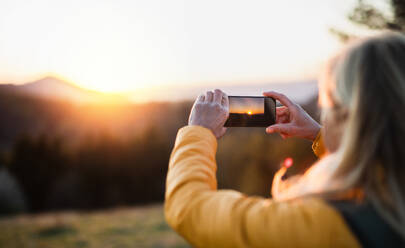Ältere Wanderin steht draußen in der Natur bei Sonnenuntergang und fotografiert mit ihrem Smartphone. - HPIF16370