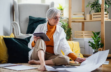 Portrait of senior woman architect sitting with blueprints on floor indoors at home, working. - HPIF16374