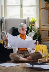 Portrait of senior woman architect sitting with blueprints on floor indoors at home, working. - HPIF16433