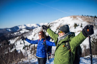 Side view of senior couple hikers with nordic walking poles in snow-covered winter nature, stretching arms. - HPIF16454