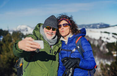 Portrait of senior couple hikers standing in snow-covered winter nature, taking selfie. - HPIF16467