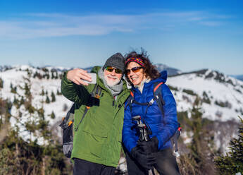 Portrait of senior couple hikers standing in snow-covered winter nature, taking selfie. - HPIF16468