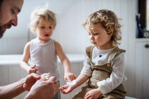 Vater mit Kindern, der einem kleinen verletzten Kleinkind zu Hause einen Verband anlegt. - HPIF16538