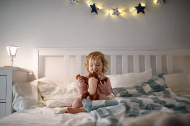 Small toddler girl sitting on bed indoors at home, playing with soft toy. - HPIF16546