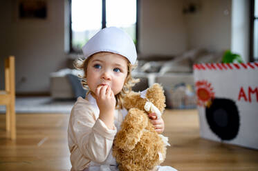 Small toddler girl with doctor uniform indoors at home, playing with teddy bear. - HPIF16567