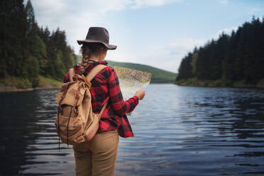 Rückansicht einer jungen Frau, die in der sommerlichen Natur am See steht und eine Karte benutzt. - HPIF16599