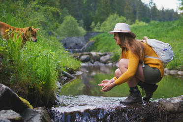 Happy young woman with dog standing by stream on a walk outdoors in summer nature, washing hands. - HPIF16612