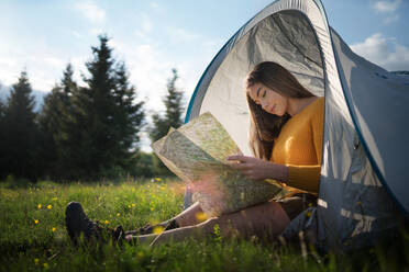 Side view of happy young woman sitting in tent shelter outdoors in summer nature, using map. - HPIF16625