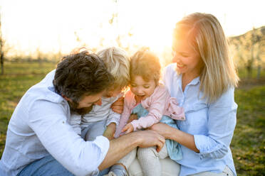 Glückliche Familie mit zwei kleinen Kindern sitzt im Frühling bei Sonnenuntergang im Obstgarten. - HPIF16762