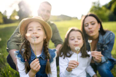 Front view of happy family with two small daughters sitting outdoors in spring nature, blowing dandelion seeds. - HPIF16793