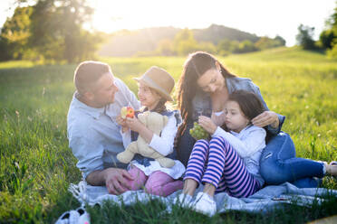 Front view of happy family with two small daughters sitting outdoors in spring nature, having picnic. - HPIF16796