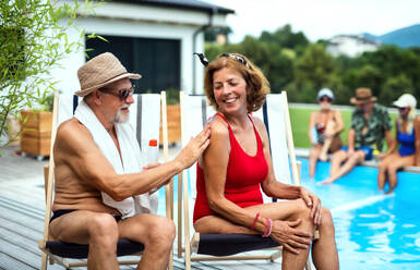 Fröhliches älteres Ehepaar sitzt am Swimmingpool im Hinterhof und benutzt Sonnencreme. - HPIF16840