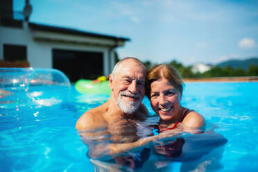 Portrait of cheerful senior couple in swimming pool outdoors in backyard, looking at camera. - HPIF16866