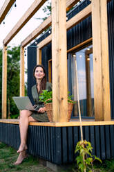 Happy young woman with laptop outdoors, weekend away in container house in countryside. - HPIF16984