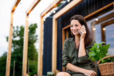 Happy young woman with smartphone outdoors, weekend away in container house in countryside. - HPIF16986