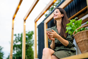Glückliche junge Frau mit Smartphone im Freien, Wochenende im Containerhaus auf dem Lande. - HPIF16989