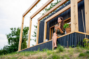 Happy young woman with smartphone outdoors, weekend away in container house in countryside. - HPIF16990