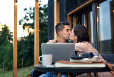 Young couple with laptop sitting and kissing outdoors, weekend away in container house in countryside. - HPIF16996