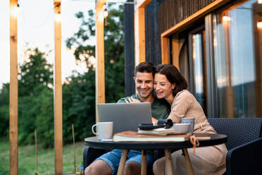 Young couple with laptop sitting outdoors, weekend away in container house in countryside. - HPIF16997