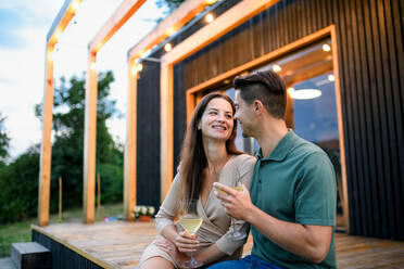 Young couple with wine sitting outdoors, weekend away in container house in countryside. - HPIF16999