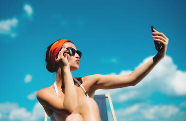 Eine junge Frau fängt einen Moment der Entspannung und des Spaßes ein, indem sie ein Selfie mit ihrem Smartphone am Swimmingpool in ihrem Garten macht - HPIF17038