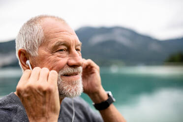 Ein älterer Mann mit Kopfhörern steht am See in der Natur und hört Musik. - HPIF17233