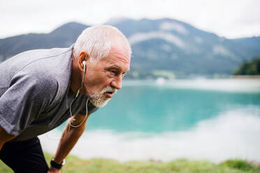 Ein älterer Mann mit Kopfhörern, der am See in der Natur steht und Sport treibt. Raum kopieren. - HPIF17234
