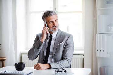 Ein seriöser, reifer Geschäftsmann mit Smartphone, der in einem Büro am Tisch sitzt und ein Telefongespräch führt. - HPIF17335