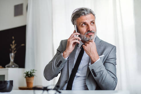 Ein seriöser, reifer Geschäftsmann mit Smartphone, der in einem Büro am Tisch sitzt und ein Telefongespräch führt. - HPIF17338