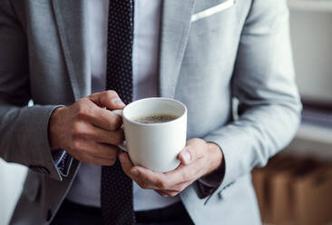 Mittelteil eines Geschäftsmannes, der in einem Büro sitzt und eine Tasse Kaffee in der Hand hält, in Großaufnahme. - HPIF17358