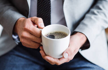 Mittelteil eines Geschäftsmannes, der in einem Büro sitzt und eine Tasse Kaffee in der Hand hält, in Großaufnahme. - HPIF17359