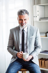 A mature businessman sitting in an office on a stool, holding a cup of coffee. - HPIF17360