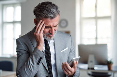Ein trauriger und frustrierter Geschäftsmann mit Smartphone steht in einem Büro und liest schlechte Nachrichten. - HPIF17369