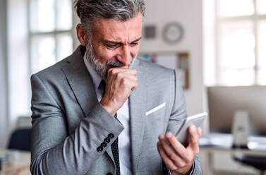 Ein trauriger und frustrierter Geschäftsmann mit Smartphone steht in einem Büro und liest schlechte Nachrichten. - HPIF17370