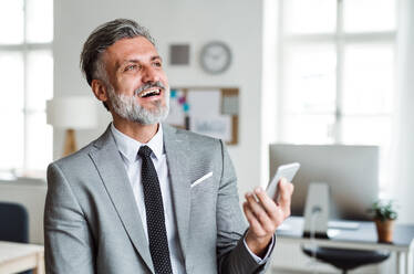 A cheerful mature businessman with smartphone standing in an office, laughing. - HPIF17372