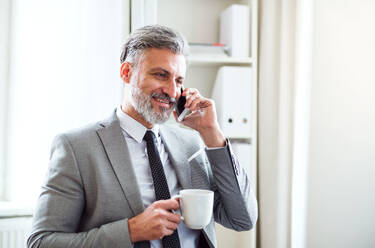 Älterer Geschäftsmann mit Smartphone und Kaffee, der in einem Büro steht und einen Anruf tätigt. - HPIF17387