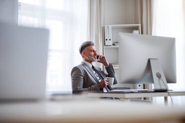 Seriöser reifer Geschäftsmann mit Smartphone, Kaffee und Desktop-Computer, der am Tisch in einem Büro sitzt und telefoniert. - HPIF17388