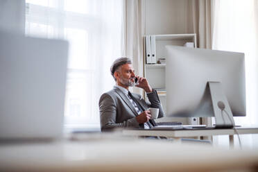 Serious mature businessman with smartphone, coffee and desktop computer sitting at the table in an office, making a phone call. - HPIF17388