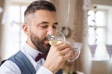 A portrait of hipster mature man indoors in a room set for a party, drinking white wine. - HPIF17416