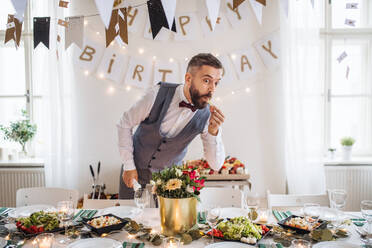 A mature man standing indoors in a room set for a party, making funny faces and nibbling at food. - HPIF17424