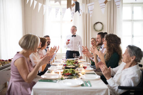 Eine große Mehr-Generationen-Familie sitzt an einem Tisch auf einer Indoor-Geburtstagsfeier, ein älterer Mann hält eine Rede. - HPIF17466