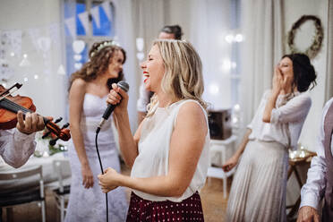 Eine junge Frau singt auf einer Hochzeitsfeier, Braut und Gäste tanzen im Hintergrund. - HPIF17537