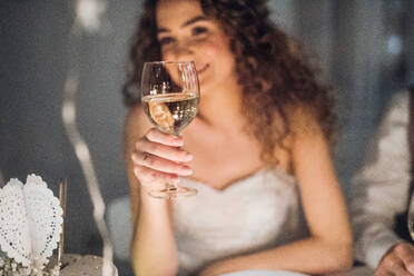 Eine Nahaufnahme einer jungen Braut, die auf einer Hochzeit an einem Tisch sitzt und ein Glas Weißwein in der Hand hält. - HPIF17554