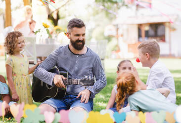 Ein Mann mit kleinen Kindern sitzt im Sommer draußen im Garten auf dem Boden und spielt Gitarre. - HPIF17586