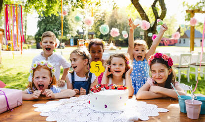 Ein Porträt von Kindern mit Kuchen, die auf einer Geburtstagsfeier im Garten im Sommer um einen Tisch herum stehen. - HPIF17644