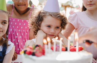 Ein Porträt von Kindern mit Kuchen, die auf einer Geburtstagsfeier im Garten im Sommer um einen Tisch herum stehen. - HPIF17645