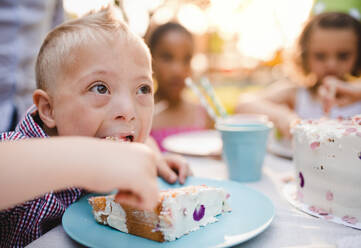 Down-Syndrom-Kind mit Freunden auf Geburtstagsfeier im Freien im Garten, Kuchen essen. - HPIF17662