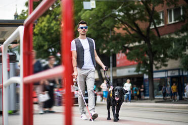 Ein junger blinder Mann mit weißem Stock und Blindenhund geht auf dem Bürgersteig in der Stadt spazieren. - HPIF17677