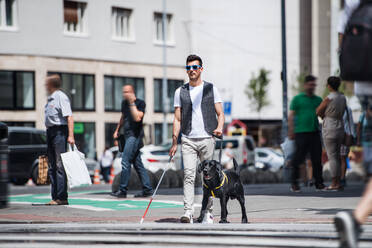 Ein junger blinder Mann mit weißem Stock und Blindenhund geht auf dem Bürgersteig in der Stadt spazieren. - HPIF17683