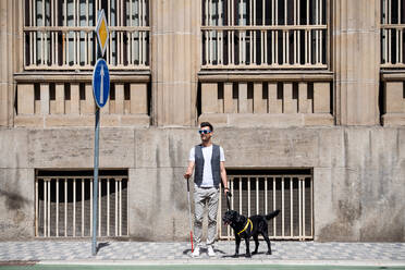 Junger blinder Mann mit weißem Stock und Blindenhund steht in der Stadt auf dem Bürgersteig und wartet. - HPIF17686